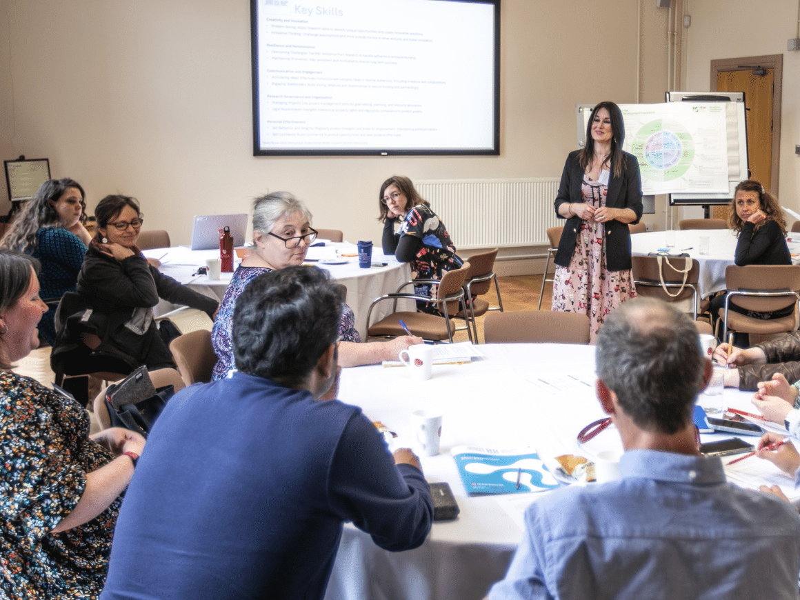 A group of early career researchers sitting round two tables, with one speaker standing up in front of a flip chart.